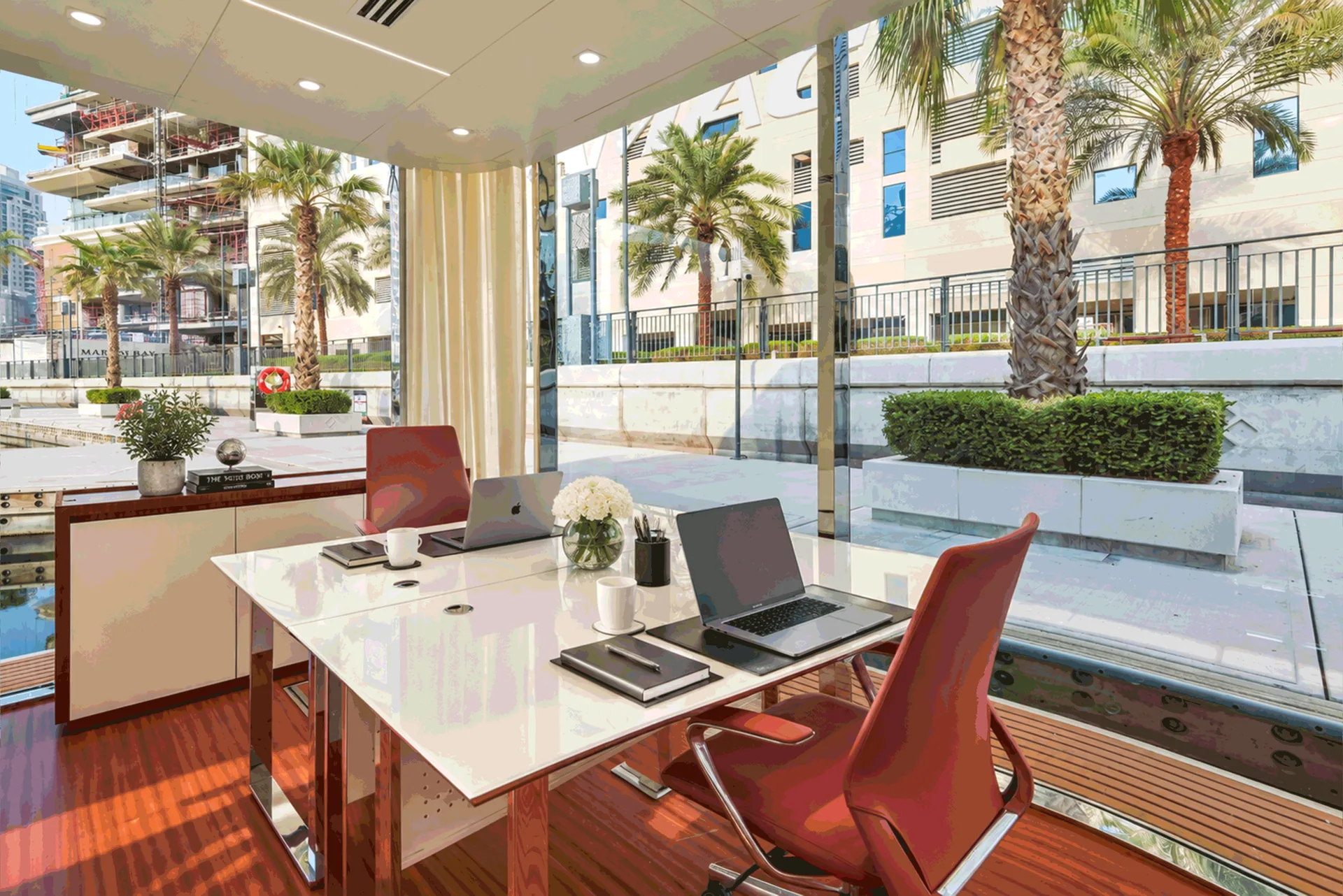 Desk with laptops beside full-height glass, palms and the quay outside