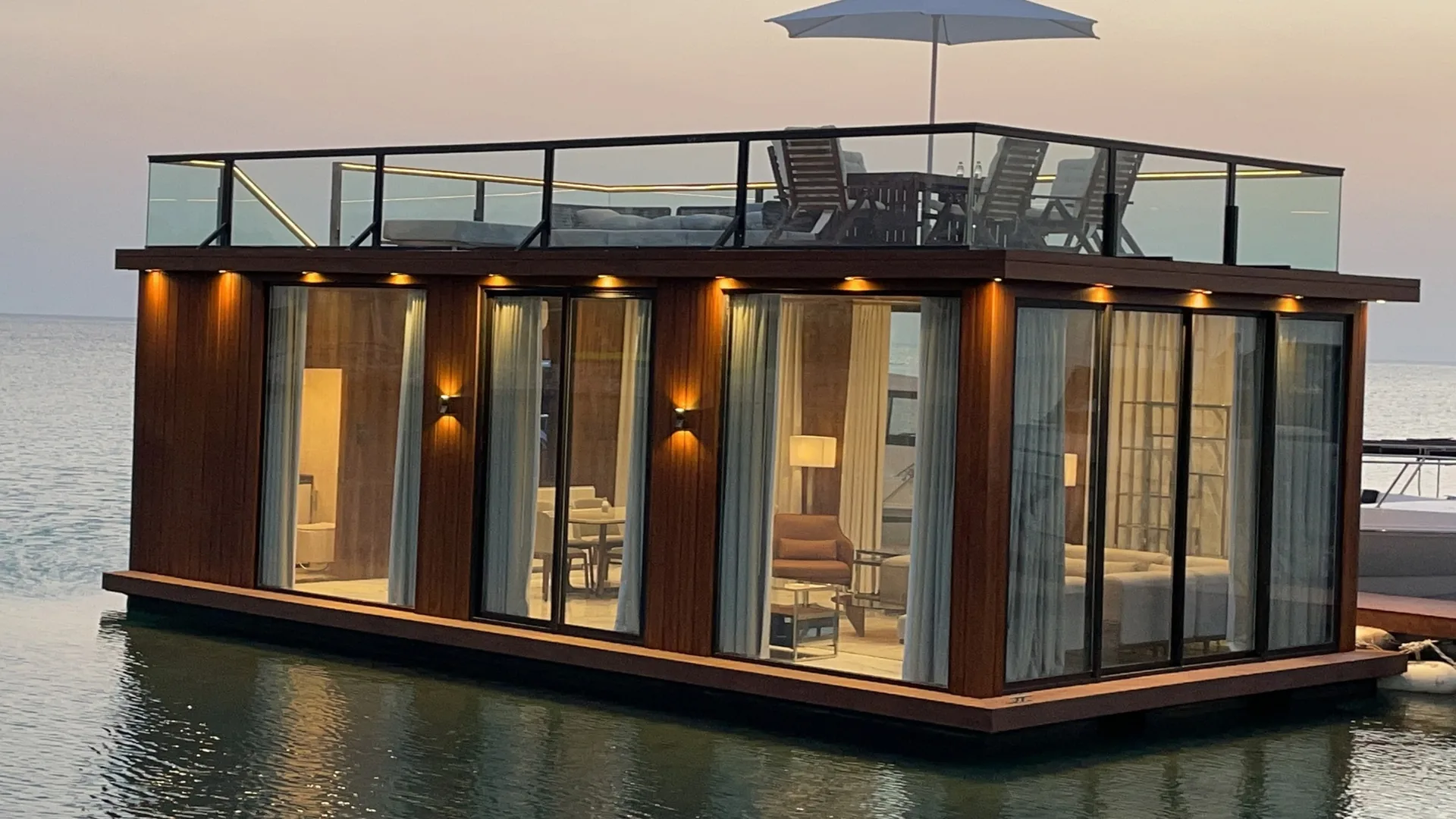 A glass-walled floating majlis lounge with a roof terrace and parasol on calm water