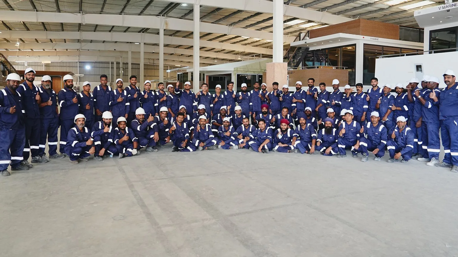 The V-Marine team in navy overalls and white hard hats, gathered for a group photo inside the shipyard
